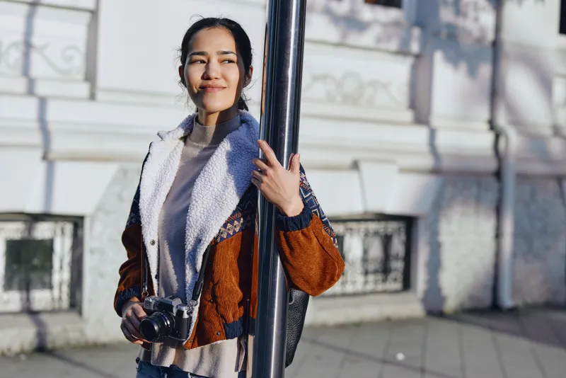Camera in hand, a casual fashion oriented woman leans by a street pole, capturing urban photography and enjoying daylight in a bright outdoor setting with a relaxed smile.