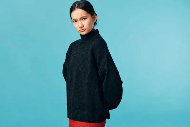 Young woman wearing black sweater stands with serious expression on blue background. Female model poses with hands behind back in casual clothing studio portrait.