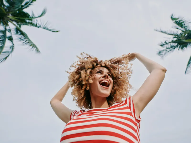 Joyful woman with curly hair wearing a red and white striped top laughs under a bright sky, captured from a low angle with palm trees framing the scene.