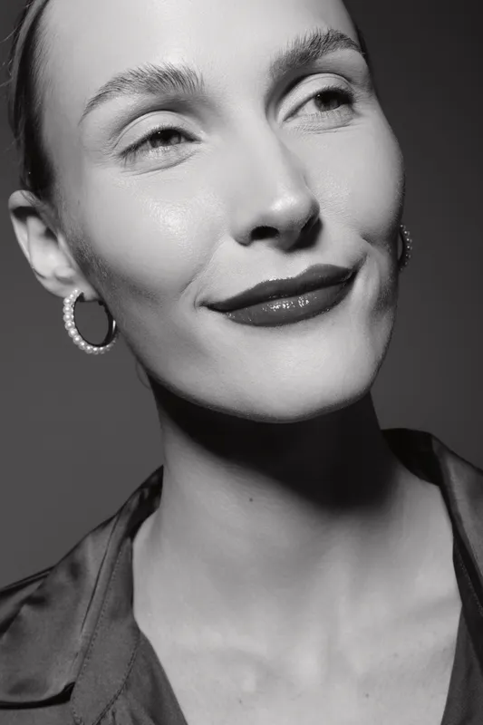 Close up of a smiling Caucasian woman with fair skin wearing pearl hoop earrings and a satin blouse. Black and white studio portrait with soft lighting and elegant mood.
