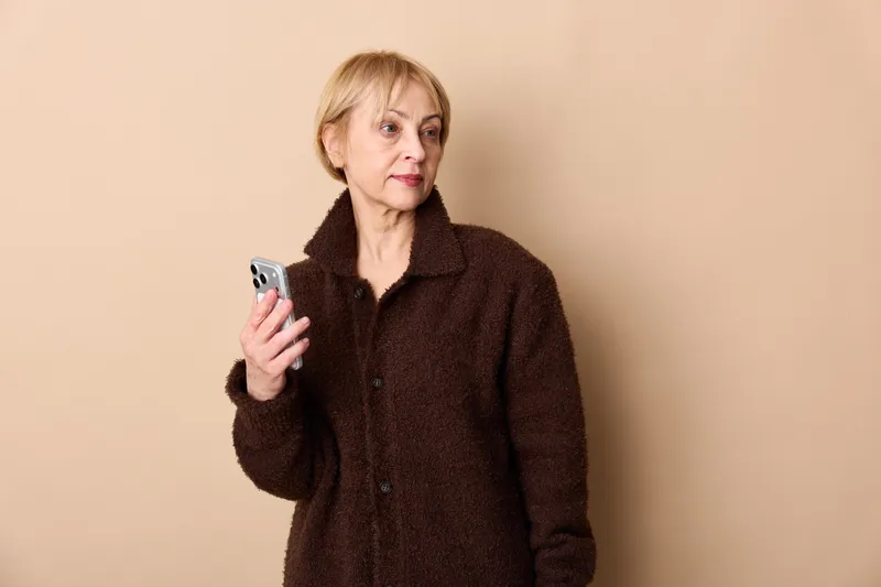 A senior woman holds a smartphone, looks worried with tense face in a warm neutral portrait indoors, concept for stress, anxiety, technology, communication, mental health