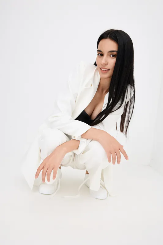 young woman squatting in white suit with long black hair, minimalistic bright background, modern style, natural makeup, casual pose, studio setting