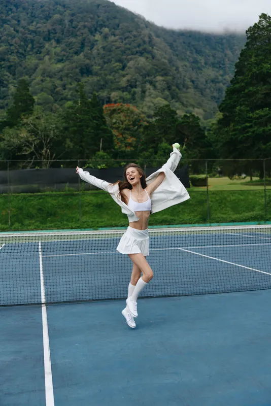 Joyful young woman celebrating on a tennis court, wearing a stylish white sports outfit, embodying a healthy lifestyle and vibrant energy against a lush green background