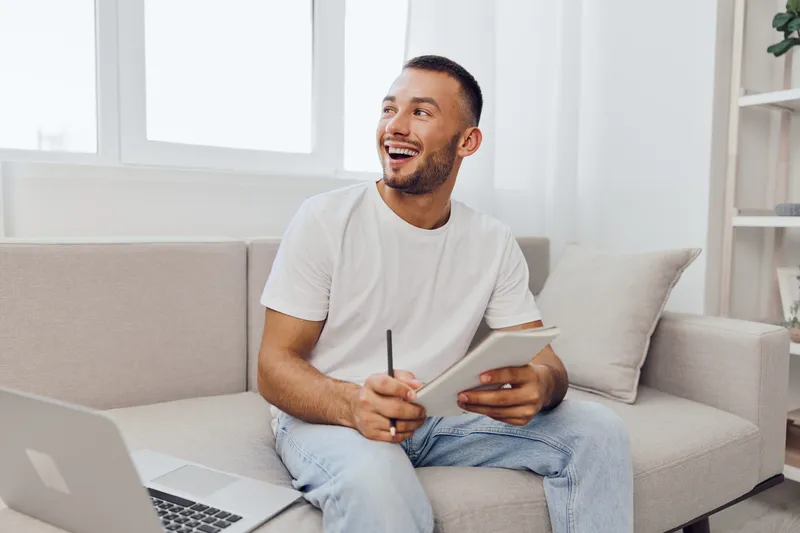 Smiling man sitting on a couch, enjoying creativity with a notepad and pen, laptop nearby, bright and modern interior setting, perfect for productivity themes.