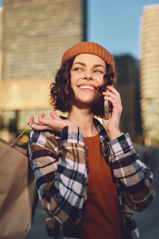 Woman shopping in city, phone to ear and smile, portrait with shopping bag and sunlight glow; authenticity and candid lifestyle captured in golden hour mood, emotional storytelling.