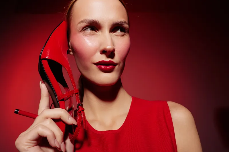 Confident woman in red dress holds red high heel shoe near face against red background. Concept of fashion, style, elegance, and bold makeup with bright lipstick and smooth skin.
