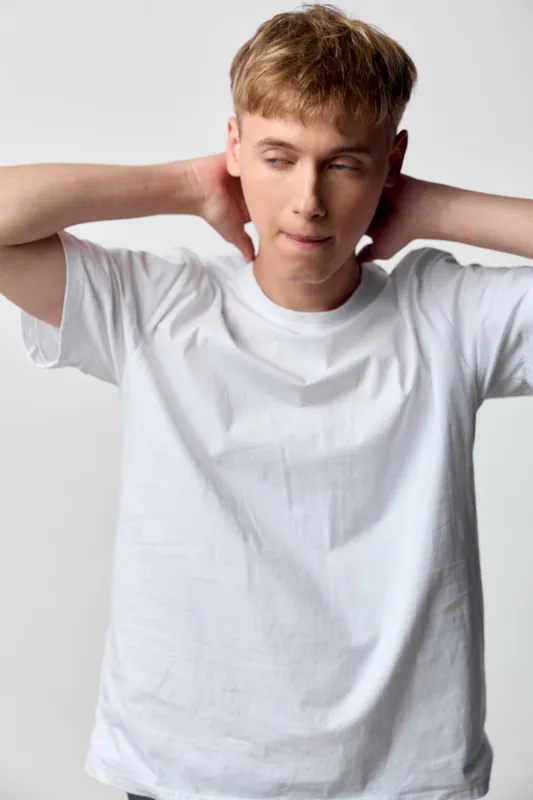 Man in white t shirt covers ears with hands, relaxed stance and concentration, casual studio setting with soft lighting and calm mood, showing a simple everyday listening moment in a minimalist space