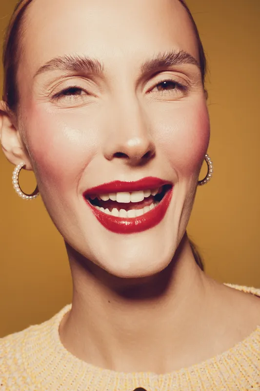 Close-up portrait of a smiling woman with red lipstick and pearl earrings on yellow background. Concept of beauty, makeup, happiness, and fashion style with natural skin and joyful expression.