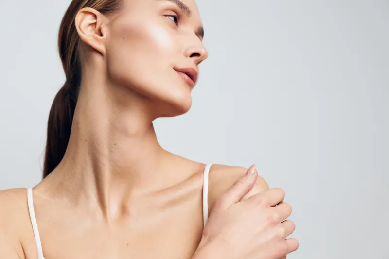 Woman portrait close up in studio light, young woman with bare shoulders, hand adjusting shoulder strap, calm expression, soft skin glow, beauty skincare routine, relaxed mood, gray background