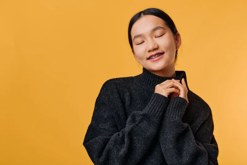 Young woman with braces wearing black sweater, eyes closed and smiling peacefully against yellow background. Portrait of calm female enjoying moment with cozy clothing and natural pose.