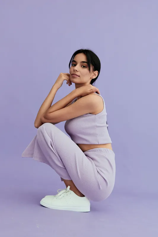 young woman squatting in casual lilac outfit and white sneakers, posing confidently on purple background with relaxed expression and natural makeup