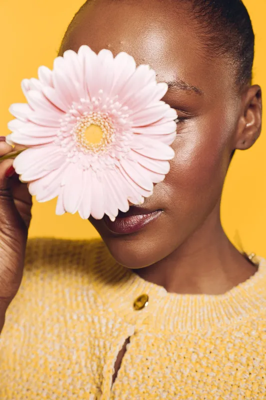 Close-up of a dark skinned African woman holding a pink flower in front of her face against a yellow background. Concept of beauty, nature, and natural skincare.