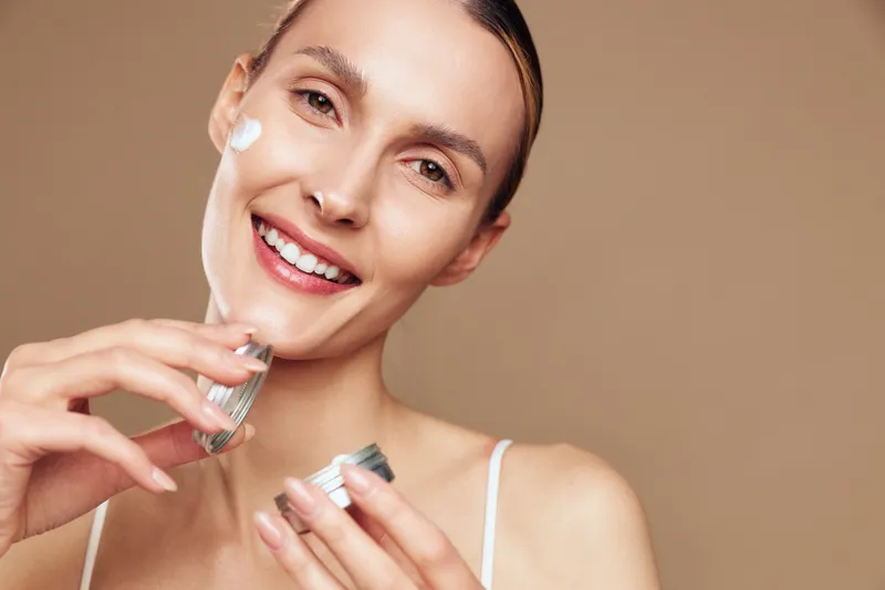 Smiling woman applying face cream with hands in close up, skincare and self care concept, warm studio lighting on brown background, soft focus.