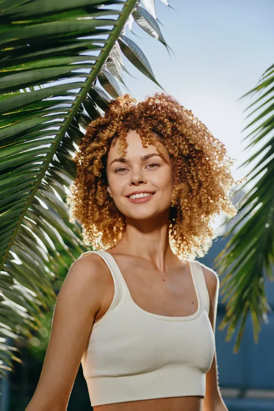 smiling woman with curly hair wearing white crop top surrounded by green palm leaves in bright natural sunlight outdoors