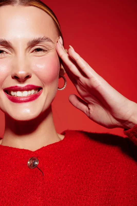 Smiling woman with red lipstick and hoop earrings poses with hand near face against red background. Close-up portrait with bright makeup and joyful expression in studio lighting.