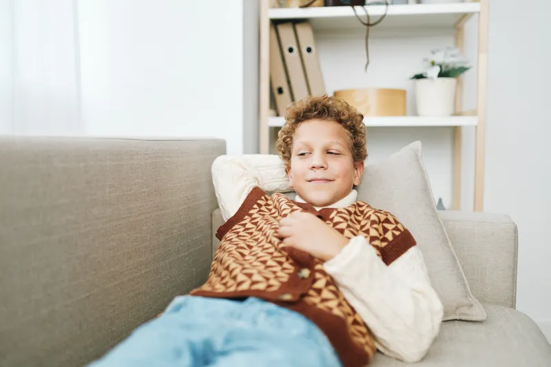 Young boy lounges on a cozy sofa, wrapped in a warm patterned blanket, relaxed and content, indoors with soft natural light and neutral decor.
