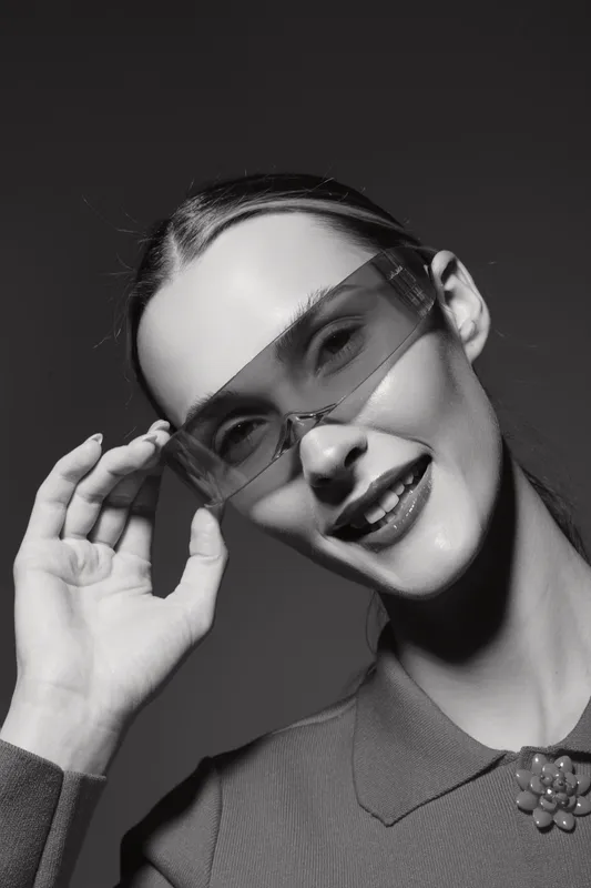 Smiling Caucasian woman with light skin wearing transparent sunglasses and a collared top adjusts glasses with hand against dark background. Fashion, portrait, style, eyewear concept.
