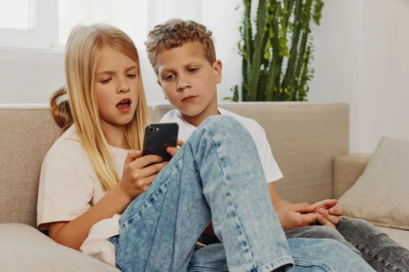 Two children sit on sofa looking at smartphone with focused expressions in bright living room. Concept of childhood, technology, and curiosity.