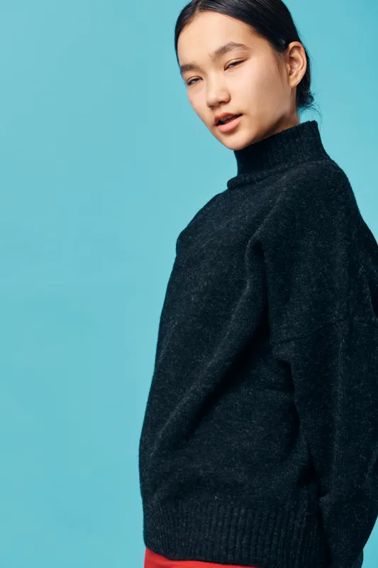 Woman in black sweater with long sleeves standing against blue background with slight smile and eyes closed. Portrait, casual outfit, young adult, relaxed pose, studio shot with soft lighting.