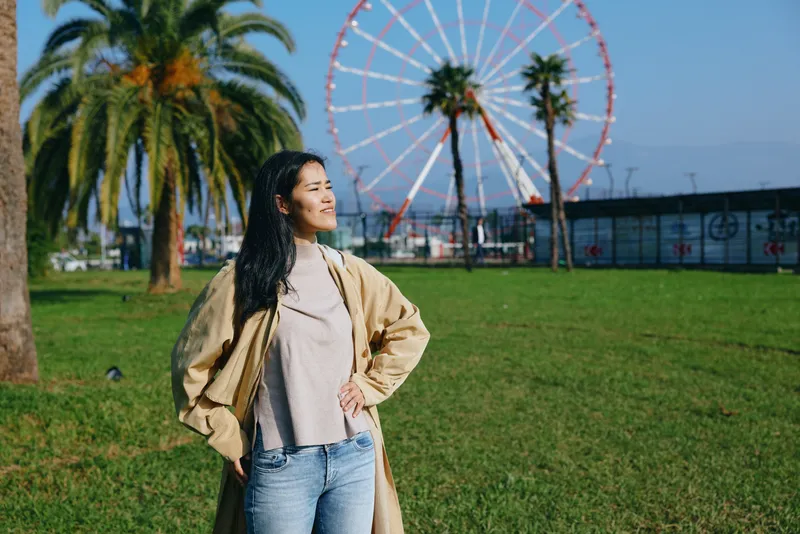 Woman in a beige cape and casual sweater stands in a sunny park with a ferris wheel in the background, enjoying outdoor leisure, relaxed pose, confident style, and fresh air.