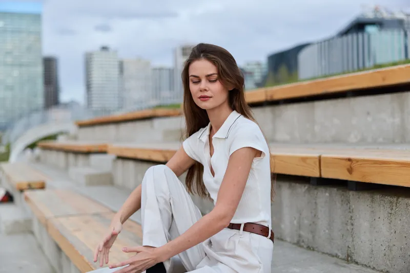 Young woman in white casual outfit sitting on urban stairs with calm expression, cityscape background, natural light, relaxed mood, spring season fashion