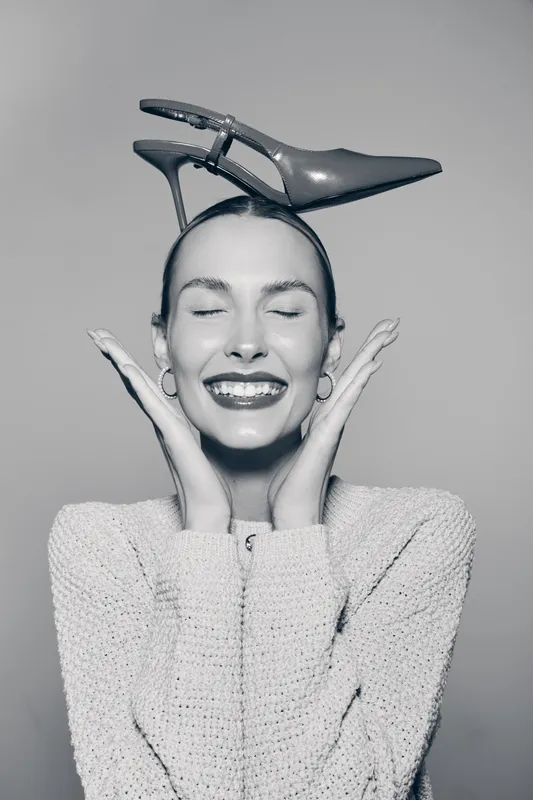Happy Caucasian woman with light skin and closed eyes holds high heel shoe on her head in studio.