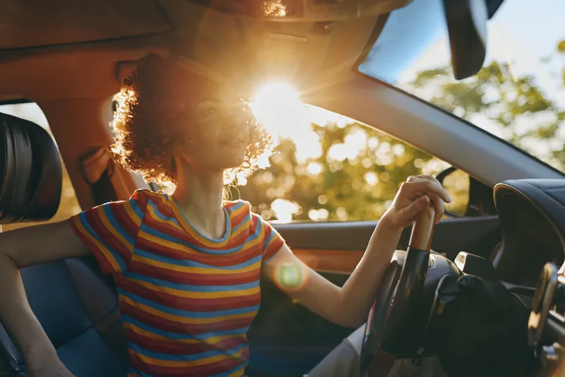 Woman driving a car in striped shirt, sunlit interior, warm retro film color, lifestyle portrait with candid motion and outdoor vibe.