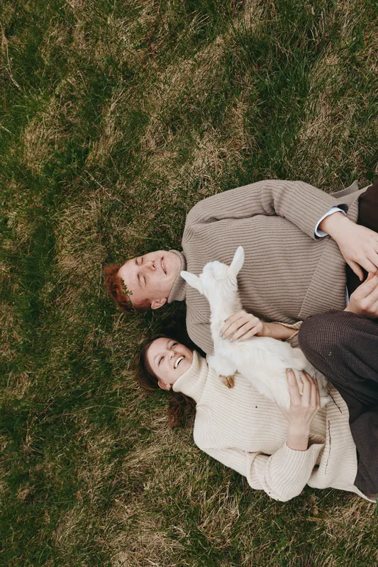 A relaxed couple lie on the grass with a playful goat kid, sharing a quiet outdoor moment. A lifestyle scene capturing affection, comfort, and a gentle bond in sunlight.