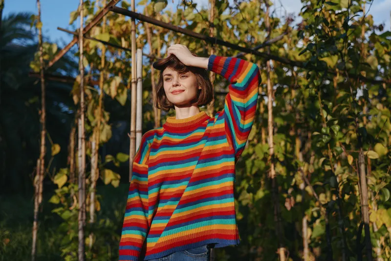 Young woman in her mid 20s wearing a striped sweater and casual outfit, short hair smiling while standing by a vine trellis in a sunlit garden, relaxed travel lifestyle portrait.