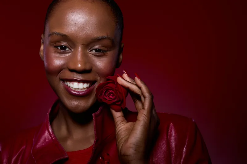 Close-up of happy African woman with dark skin holding red rose near face. She smiles warmly wearing red leather jacket against red background. Concept of beauty, joy, and self-expression.