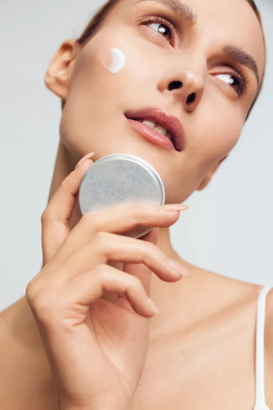 Skincare, woman, face, closeup, applying cream on cheek, holding cosmetic mirror, smooth skin, studio white background, soft light, gentle expression, beauty care routine, minimal makeup