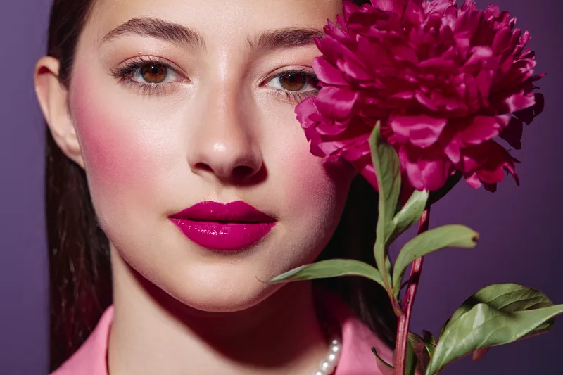 Close-up of a beautiful woman with glossy pink lipstick holding a pink peony flower, wearing pearl necklace and light pink blouse on purple background. Beauty concept.