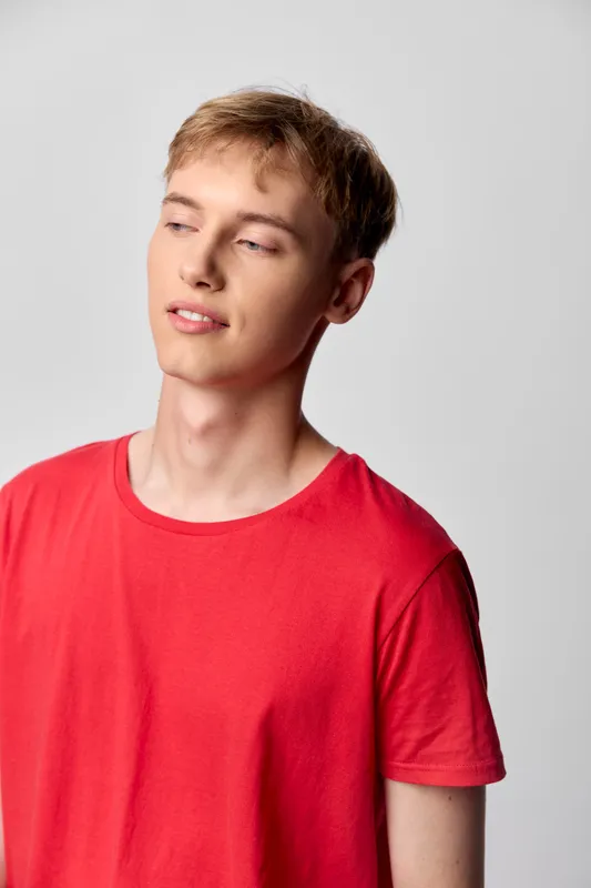 Young man in a red shirt stands against a neutral backdrop with a contemplative, relaxed gaze, conveying casual confidence in a clean studio setting for lifestyle and fashion projects