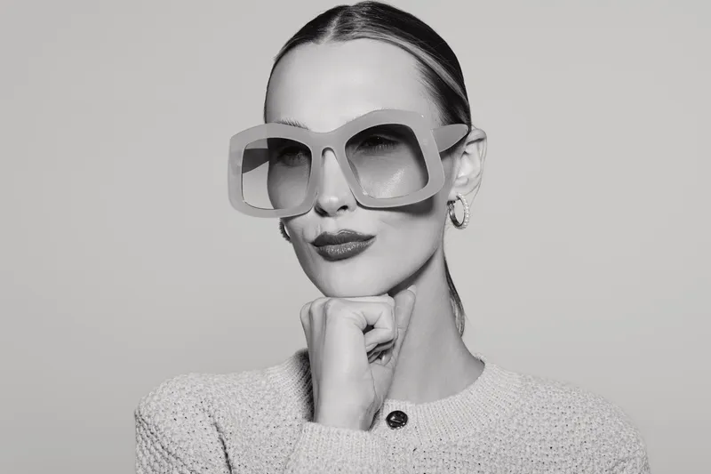 Stylish Caucasian woman with pale skin wearing oversized sunglasses poses with hand under chin. Black and white studio portrait with minimalist fashion concept and confident expression.
