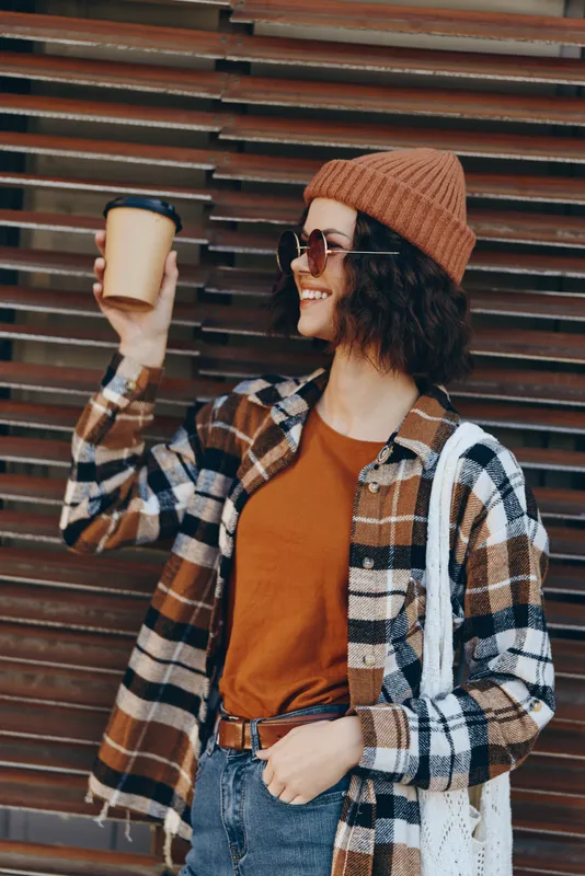 Woman holding coffee with a smile in urban streetwear wearing beanie, sunglasses and plaid jacket with denim. Authenticity, candid lifestyle, golden hour glow and emotional storytelling mood.