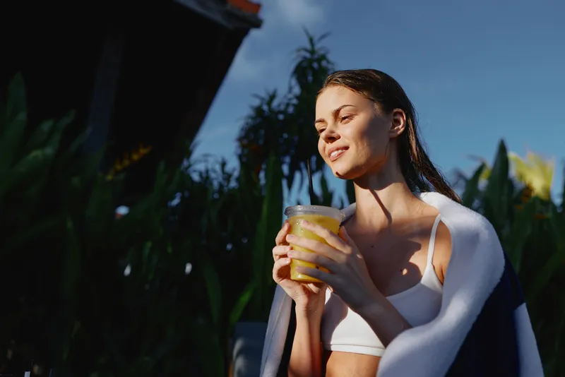 Cheerful woman outdoors, enjoying a warm beverage in the sun, relaxed posture, natural colors, lush greenery background, casual summer moment, vibrant mood, lifestyle oriented