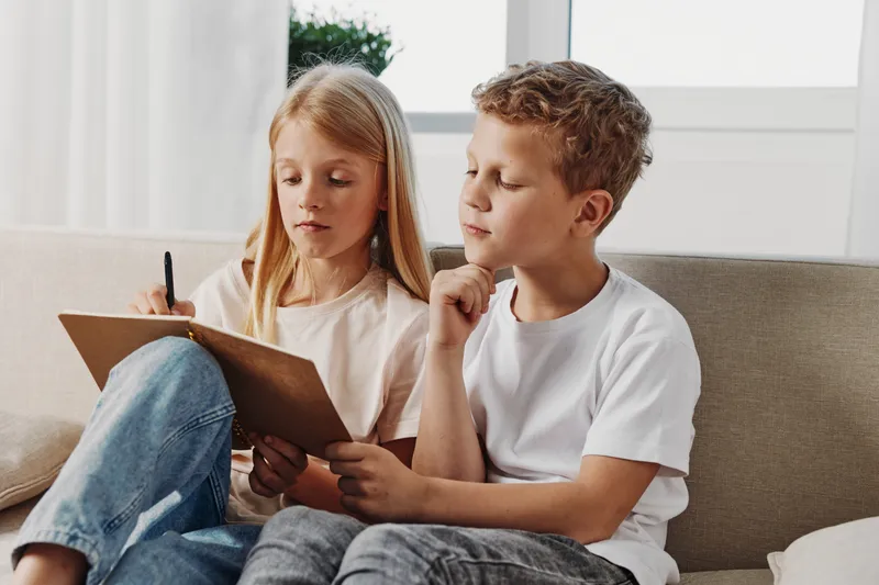 Two children sitting on a couch focused on writing and reading on a clipboard in a bright room. Concept of learning, education, concentration, and childhood development.