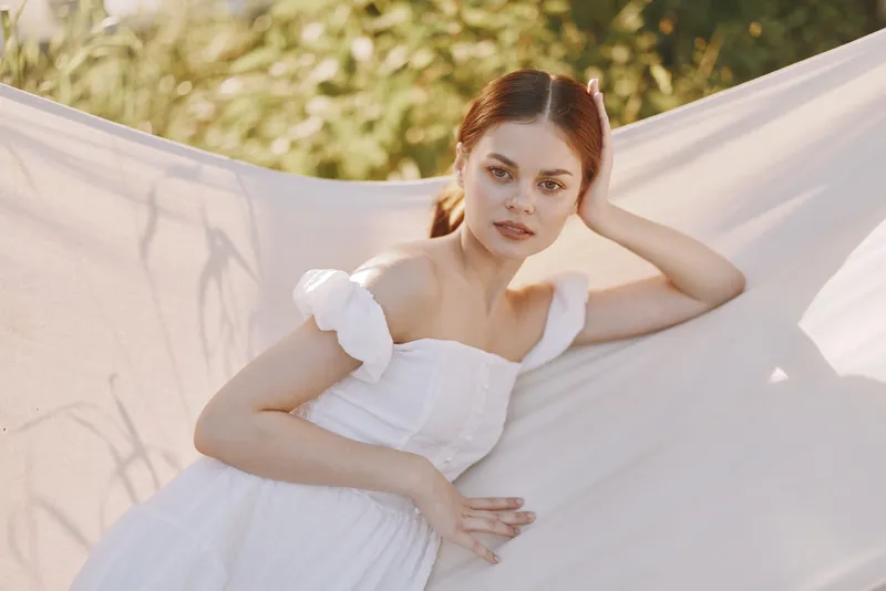 A serene young woman wearing a white dress poses gracefully against a soft natural backdrop, evoking tranquility and elegance in a bright, sunny setting