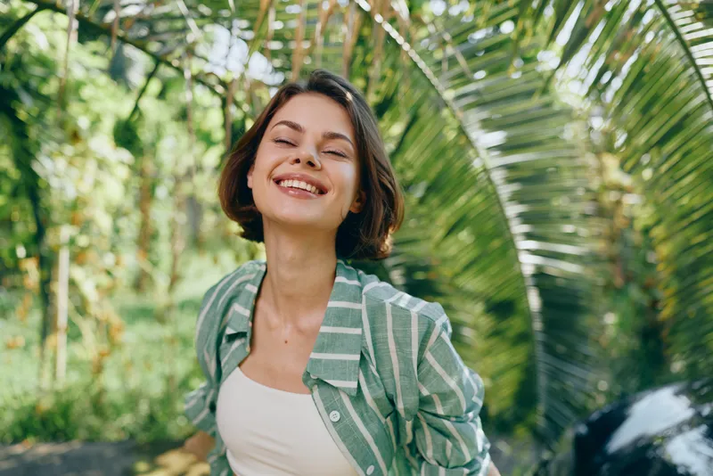 A joyful woman stands outdoors among lush greenery, smiling warmly at the camera. Casual jacket, sunny day, natural light highlights her confident, friendly presence.