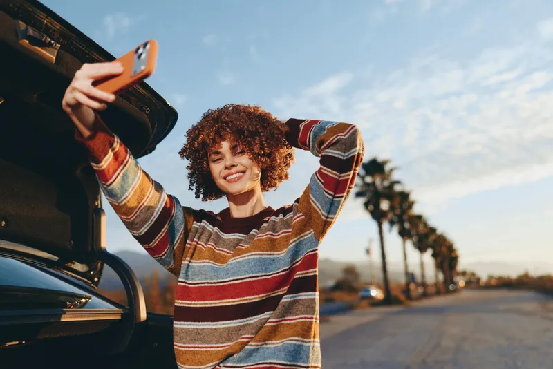 Woman lifestyle outdoors, smiling curly hair, holding smartphone by an open car, warm sunlit road, relaxed happy pose, casual striped sweater, palm trees blurred background daylight