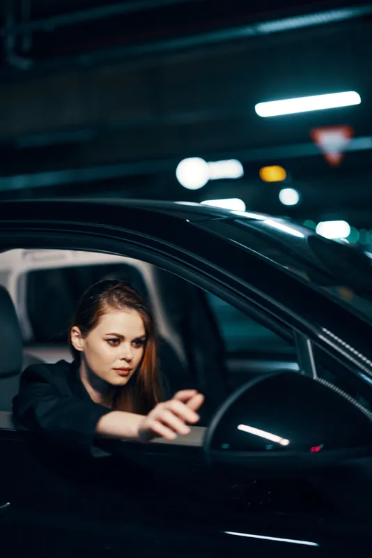 Вертикальный снимок сбоку, ночь: женщина сидит в автомобиле, смотрит в окно и тянется рукой к боковому зеркалу заднего вида