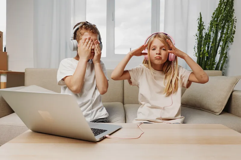 Two children with headphones sit on a couch using a laptop, showing playful and focused moods in a bright living room.