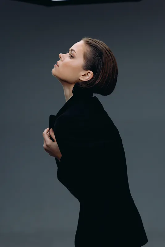 Profile of a woman in black coat with closed eyes, styled hair, and calm expression on gray background.