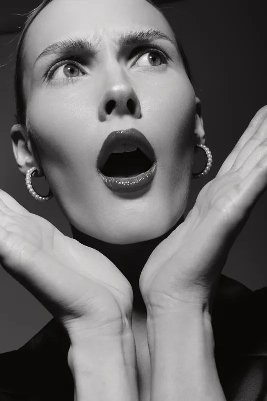 Close-up of a surprised white woman with pale skin and dark lipstick, hands framing her face. Studio portrait in black and white with dramatic lighting and hoop earrings.