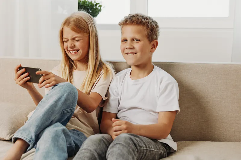 Two happy children sitting on sofa playing video game together, enjoying leisure time in bright living room.