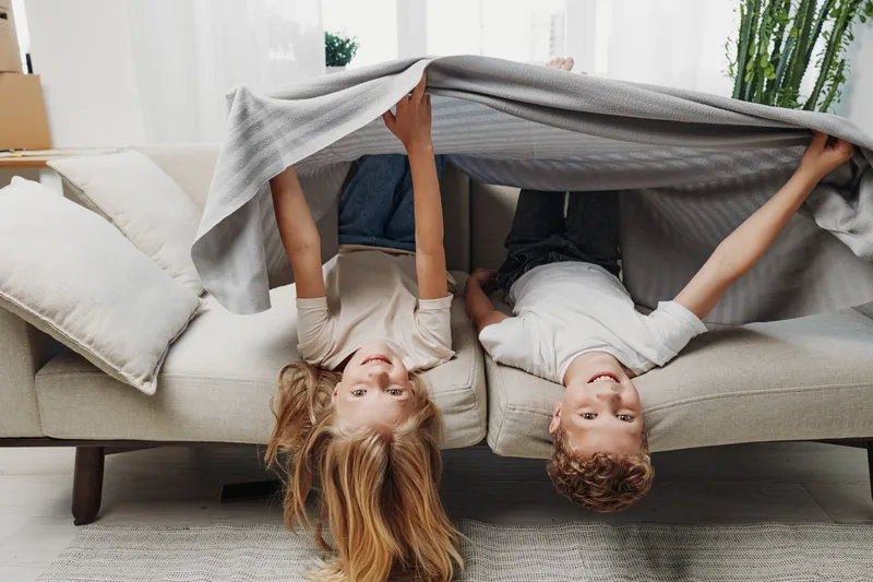Two happy children lying upside down on a sofa covered with a blanket in a bright living room. Concept for childhood fun, family moments, and cozy home atmosphere.
