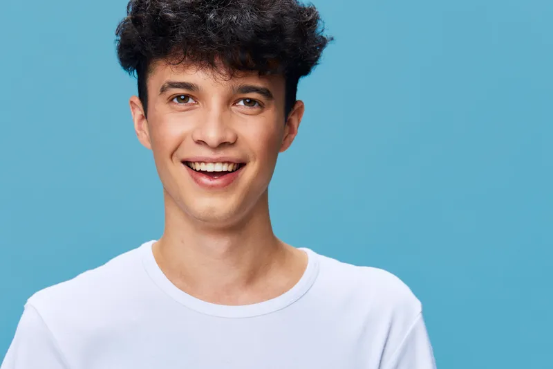 cheerful young man with curly hair smiling brightly against a blue background, wearing a casual white t-shirt, conveying happiness and positivity.