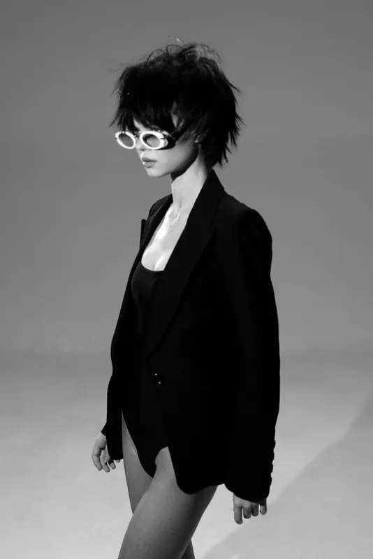 Black and white portrait of a stylish woman wearing a blazer and sunglasses in a minimalistic studio setting.