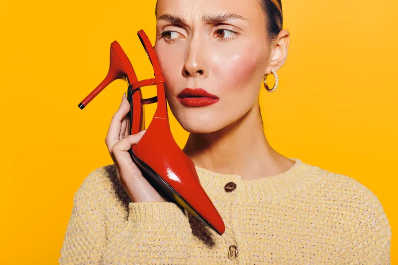 Young woman with red lipstick holds red high heel shoe near face, wearing beige sweater on bright yellow background. Concept of fashion, style, and beauty accessories.