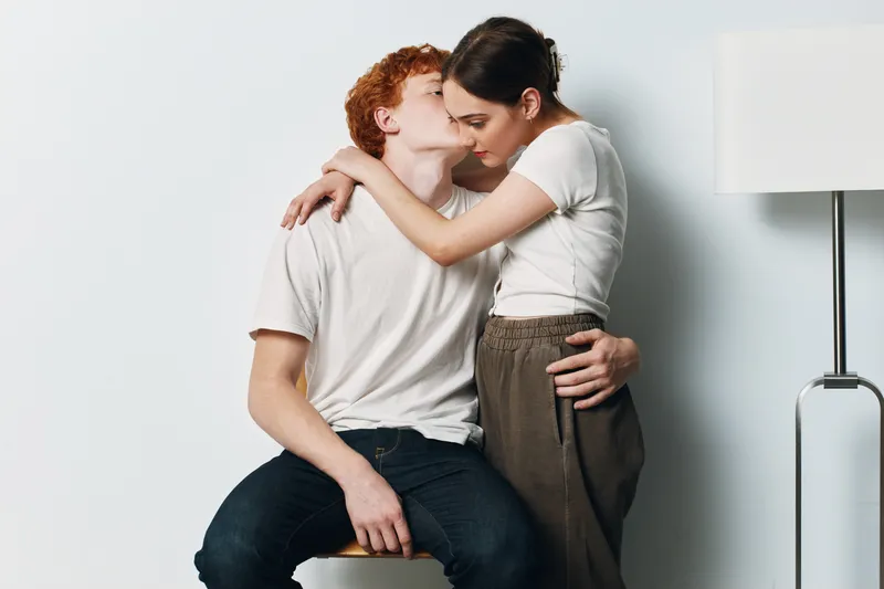 Couple, love, affection, kiss and hug of man and woman in casual clothes at home with white wall and lamp for relationship, intimacy and romance.